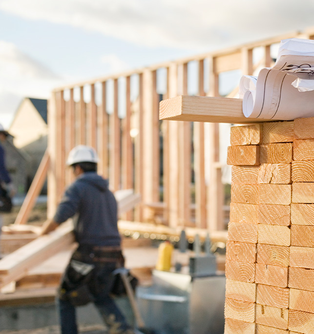 Image of a new home construction site with blueprints sitting on a stack of 2x4s and a construction worker in the background.
