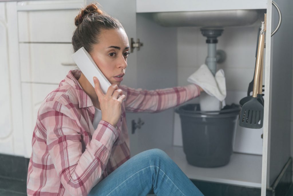 Photo of a woman wearing a plaid shirt and jeans, sitting on the floor of her kitchen, with her sink cabinet open that is leaking while she is on the phone.