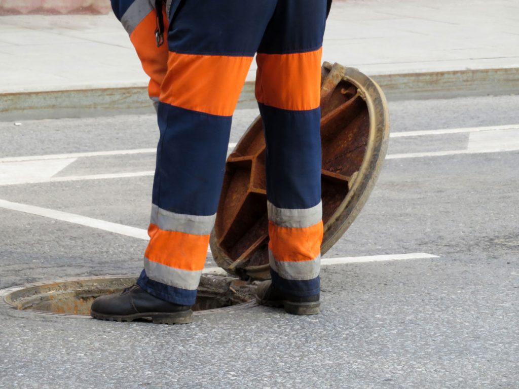 Photo of a city sewer drain being opened by a person in construction safety clothing.