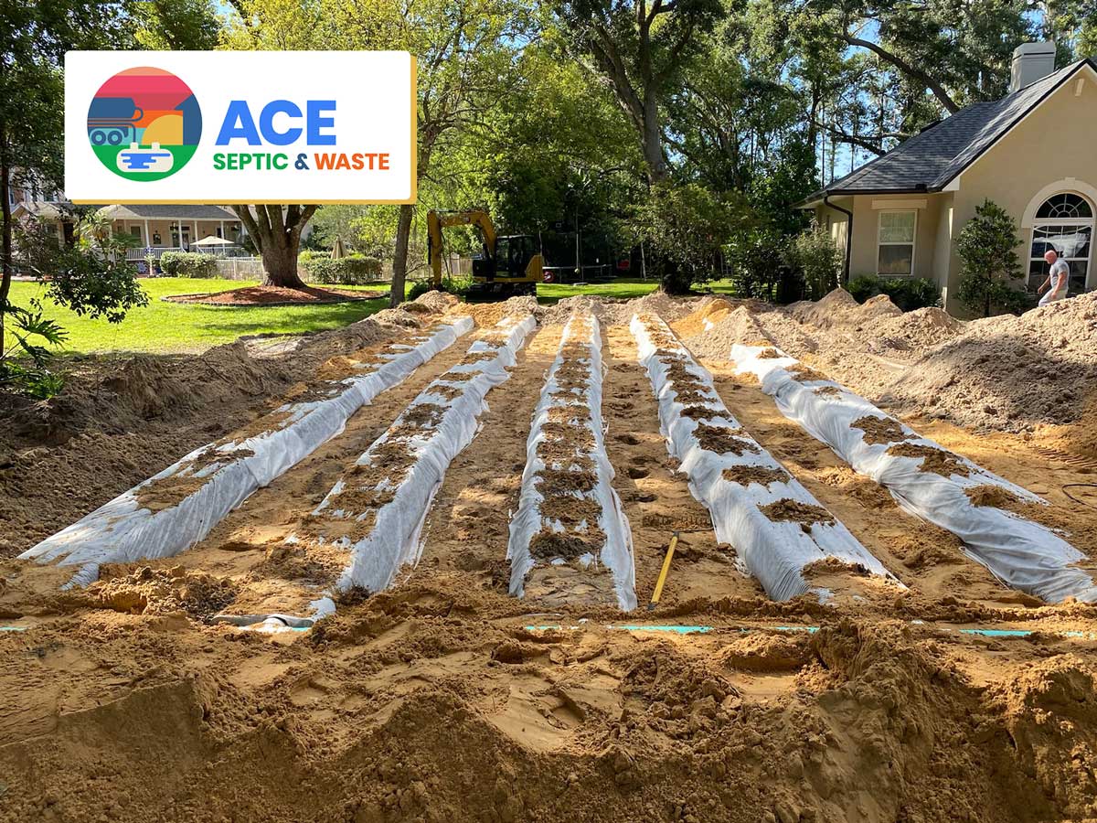 Photograph of a sunny day in Florida in the tree lined front yard  small, new beige house. The front yard ground is all dug up and 5 long pipes covered in plastic sit in the ground as part of a septic mound system.