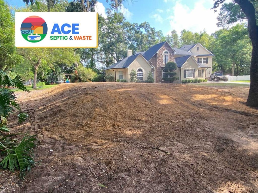 Photograph of a sunny day in Florida in the tree lined front yard of a large, new beige and bricj house. The front yard shows a shallow but visible mound covered in dirt, which is a newly installed septic mound system which was done by ACE Septic & Waste.