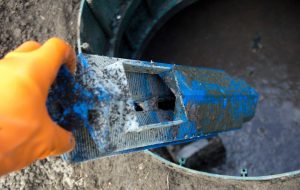 Photograph of a closeup of an orange gloved hand reaching into a circular opening to a septic system and removing a blue slatted plastic filter covered in detris.