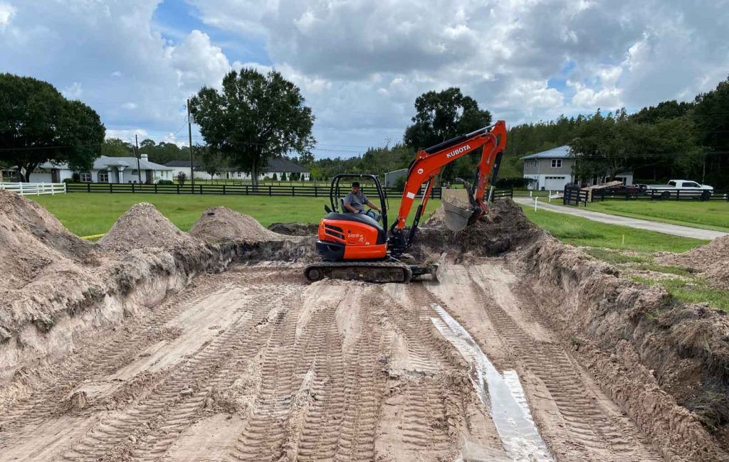 Photograph of blue blue cloudy sky, tall green trees, and suburban Florida houses in the background. In the foreground is a red digger excavator machine digging into a flat ground surface, surrounded by piles of dirt on the edges.