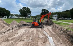 Photograph of blue blue cloudy sky, tall green trees, and suburban Florida houses in the background. In the foreground is a red digger excavator machine digging into a flat ground surface, surrounded by piles of dirt on the edges.