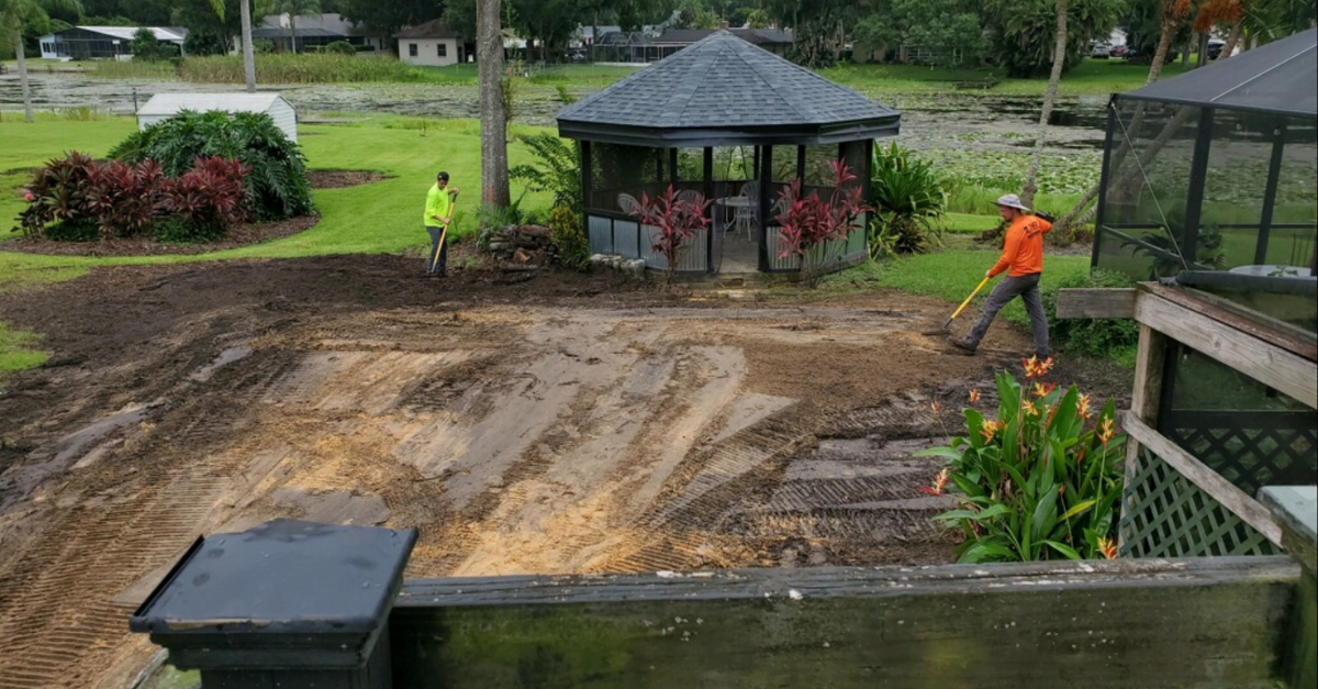 Photograph taken from a porch looking down into a grassy yard with a large portion being brown dirt. In front of a small gazebo, two people in safety color neon t-shirts smooth the dirt with rakes.