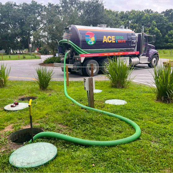 Photograph of green trees, green lawn, and gray paved road. A large purple septic pumping truck is parked with a green hose connected from its tank to a septic tank hole.