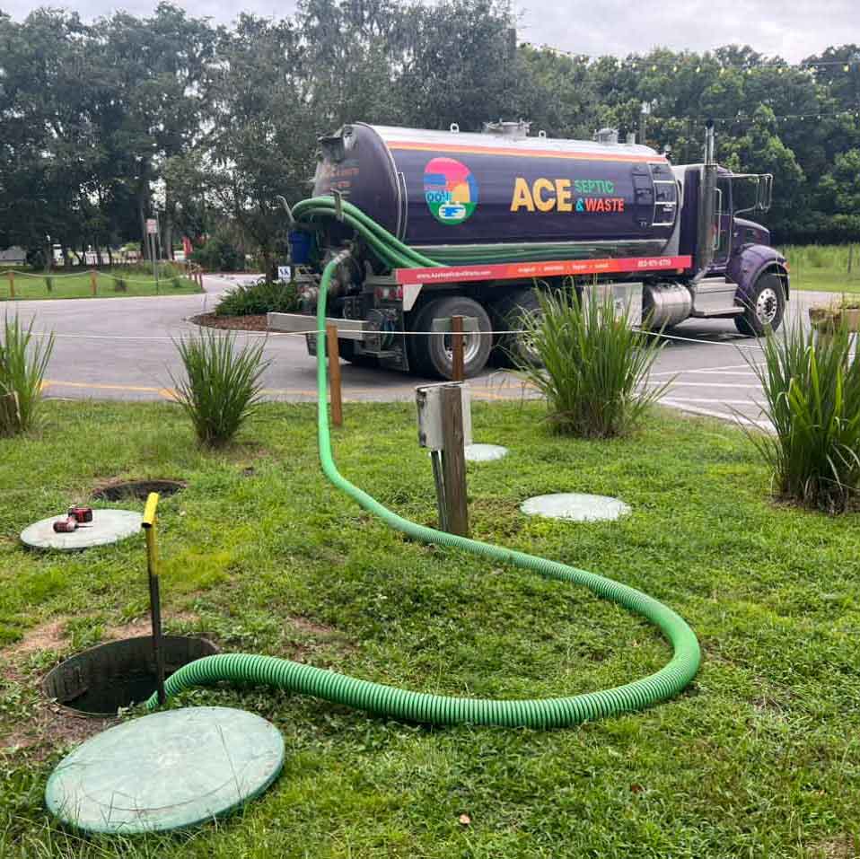 Photograph of green trees, green lawn, and gray paved road. A large purple septic pumping truck is parked with a green hose connected from its tank to a septic tank hole.