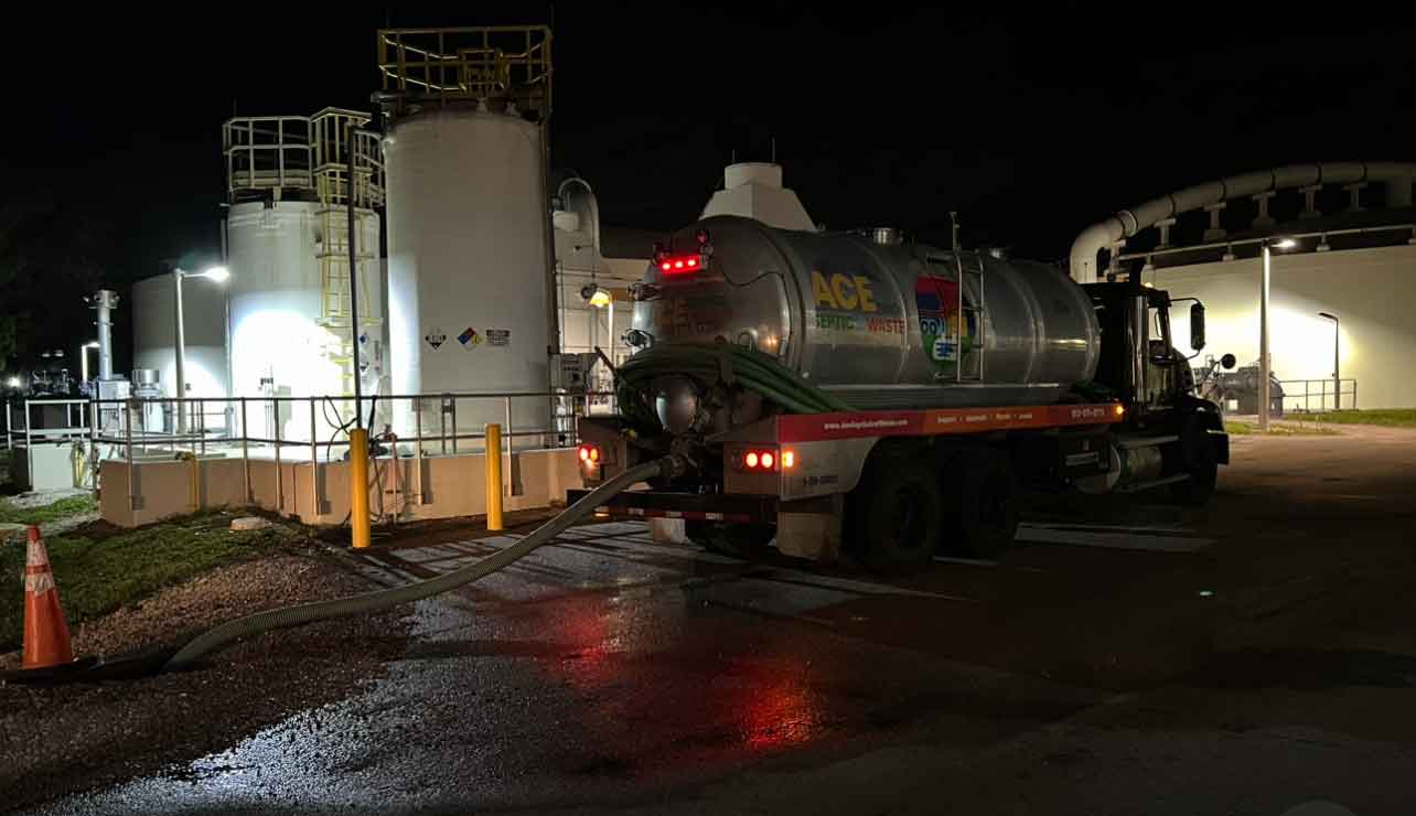Photo of a black night sky and a purple + mirror finish large commercial septic pump trunk in front of a large commercial septic system behind a fence.
