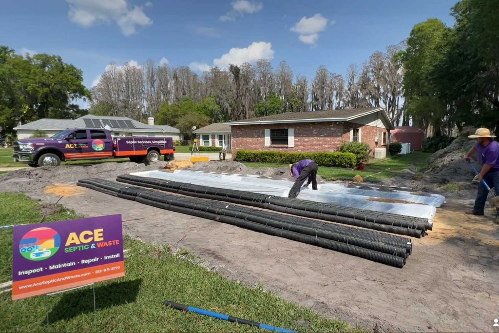 Photograph of a large side yard of a brown ranch home in Florida. A purple Dodge Ram truck with a decal that says "ACE Septic & Waste" sits in the back. Large areas have been dug out, and two works are setting up septic system components.