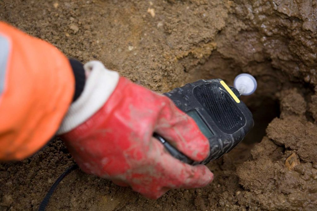 Photograph of a close up of a gloved red hand holding an electronic device that is small and used for soil testing. The device is reading a large mound of dirt.