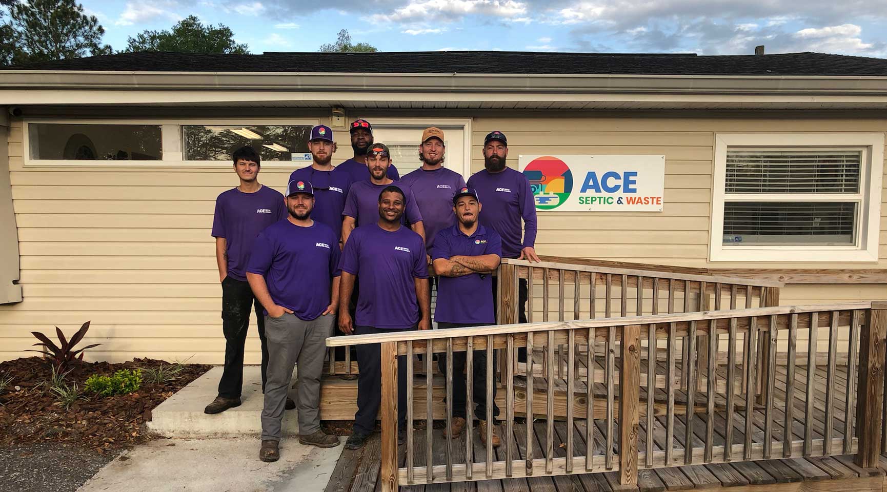 Photo of 9 people in purple shirts in front of a small office building with the ACE Septic & Waste sign on it.