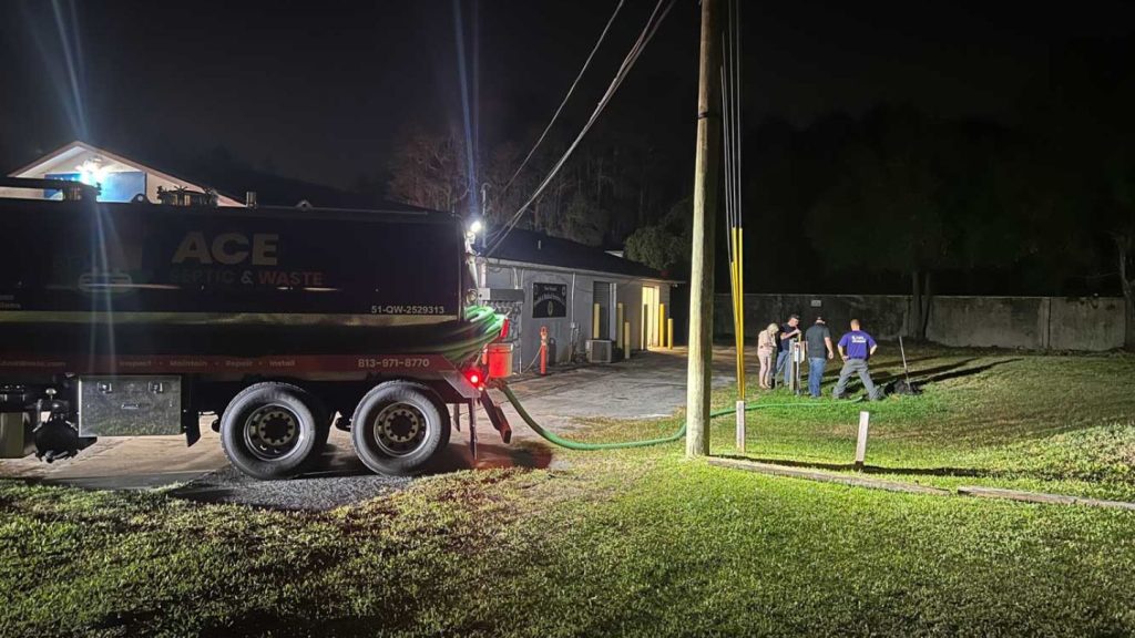 Photograph of a septic truck at night outside of a business. A group of four people inspect the ground.